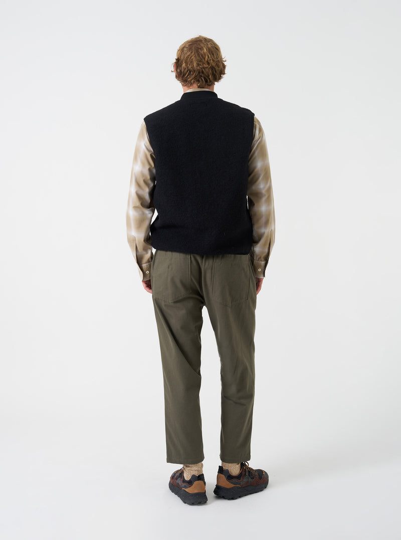 A male model showcases the Universal Works Black Wool Fleece Zip Waistcoat worn unzipped over layers against a neutral studio background. The image highlights the relaxed silhouette, open front styling, and textured fleece material.