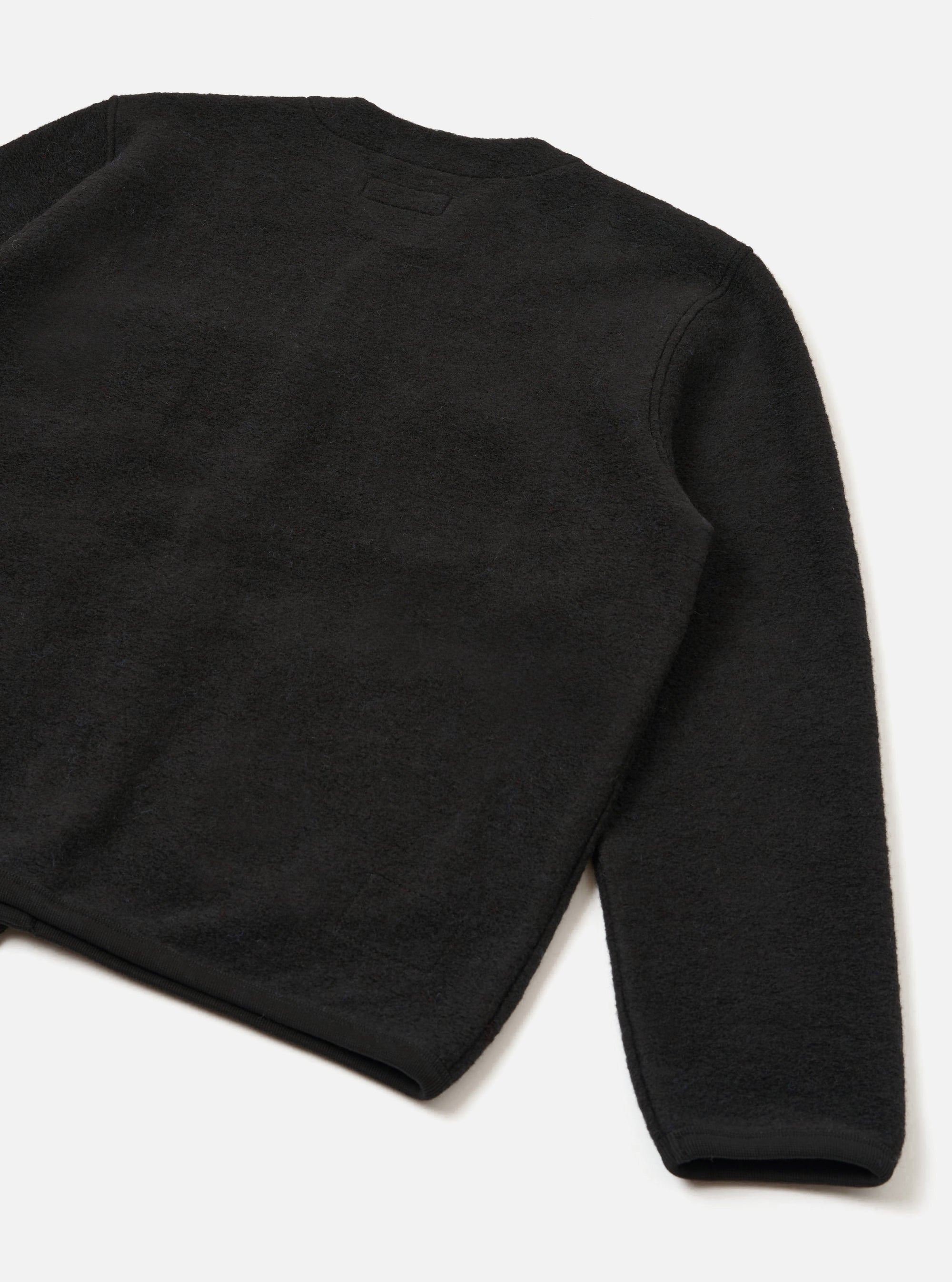 Flat lay of a black wool fleece cardigan on a white studio surface, shot from behind at an angle. The textured, fluffy fleece fabric is visible across the back panel and one full sleeve. A small tonal label sits at the upper back. The ribbed cuff is visible at the sleeve hem.