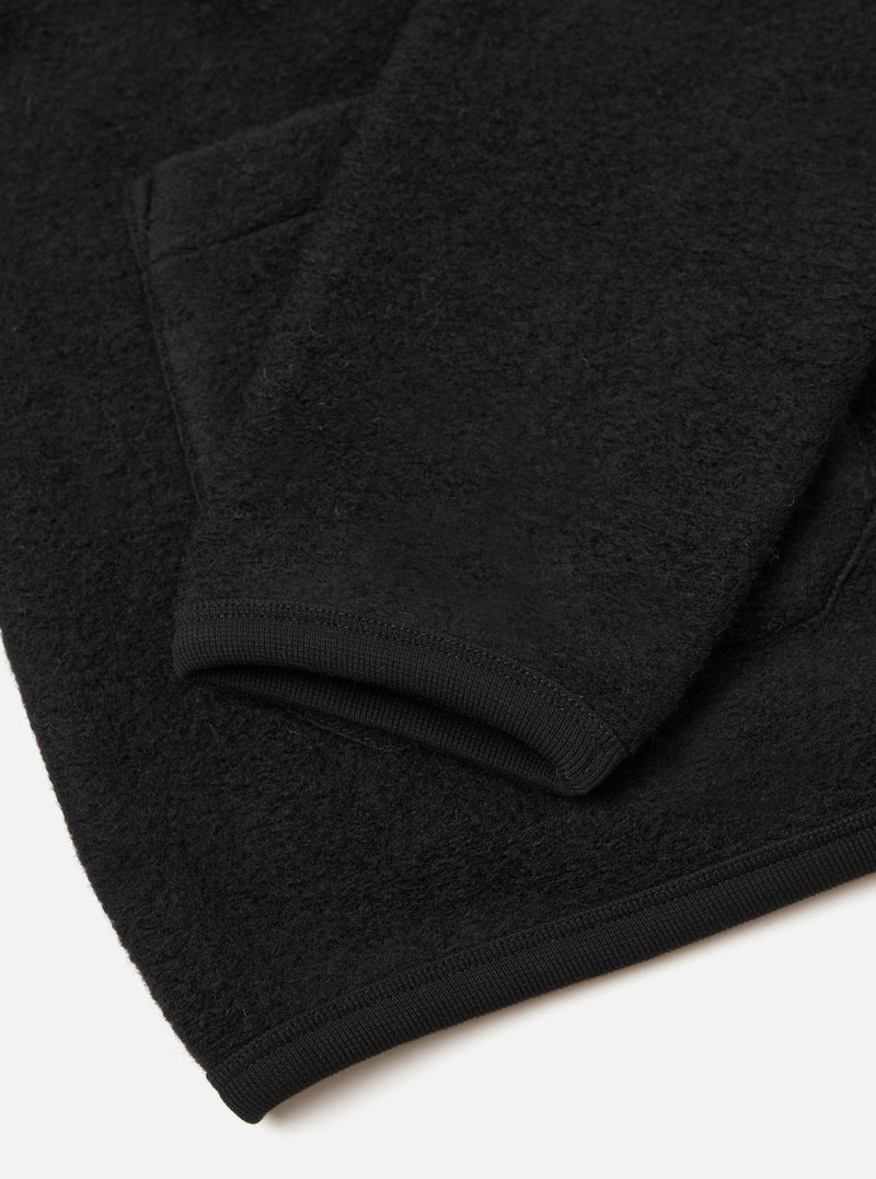 Close-up flat lay of a black wool fleece cardigan showing the sleeve cuff and lower hem. The fabric has a dense, textured fleece surface. The ribbed cuff is smooth and fitted, finished with visible stitching. The hem carries the same ribbed trim. Shot on a white studio surface.