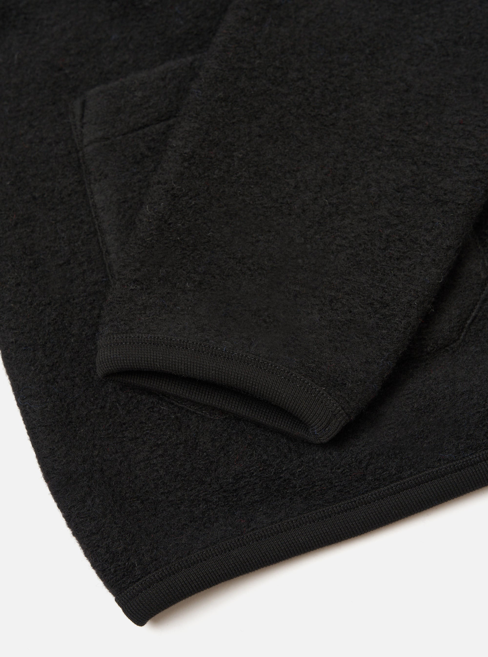 Close-up flat lay of a black wool fleece cardigan showing the sleeve cuff and lower hem. The fabric has a dense, textured fleece surface. The ribbed cuff is smooth and fitted, finished with visible stitching. The hem carries the same ribbed trim. Shot on a white studio surface.
