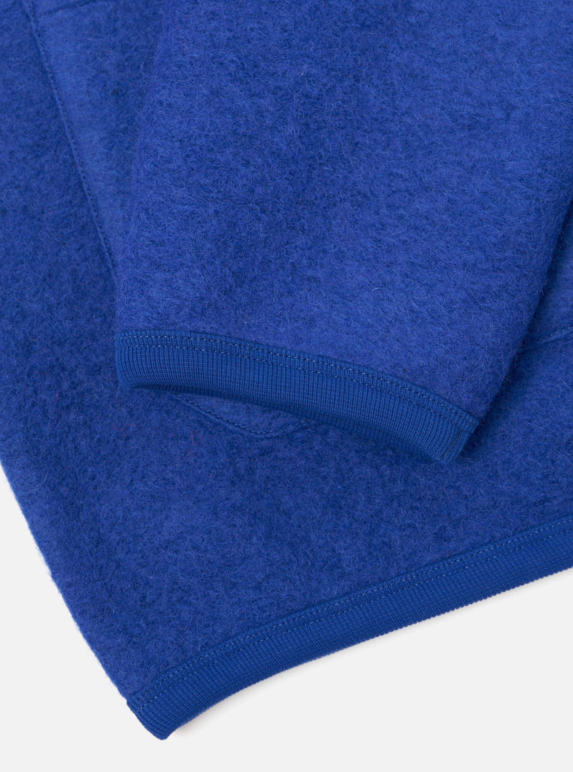 Close-up detail of a blue wool fleece cardigan laid flat on a white surface, showing the lower sleeve and hem. The boiled wool texture is clearly visible. Both the ribbed cuff and ribbed hem trim are shown in matching royal blue, with neat topstitching along the seams.