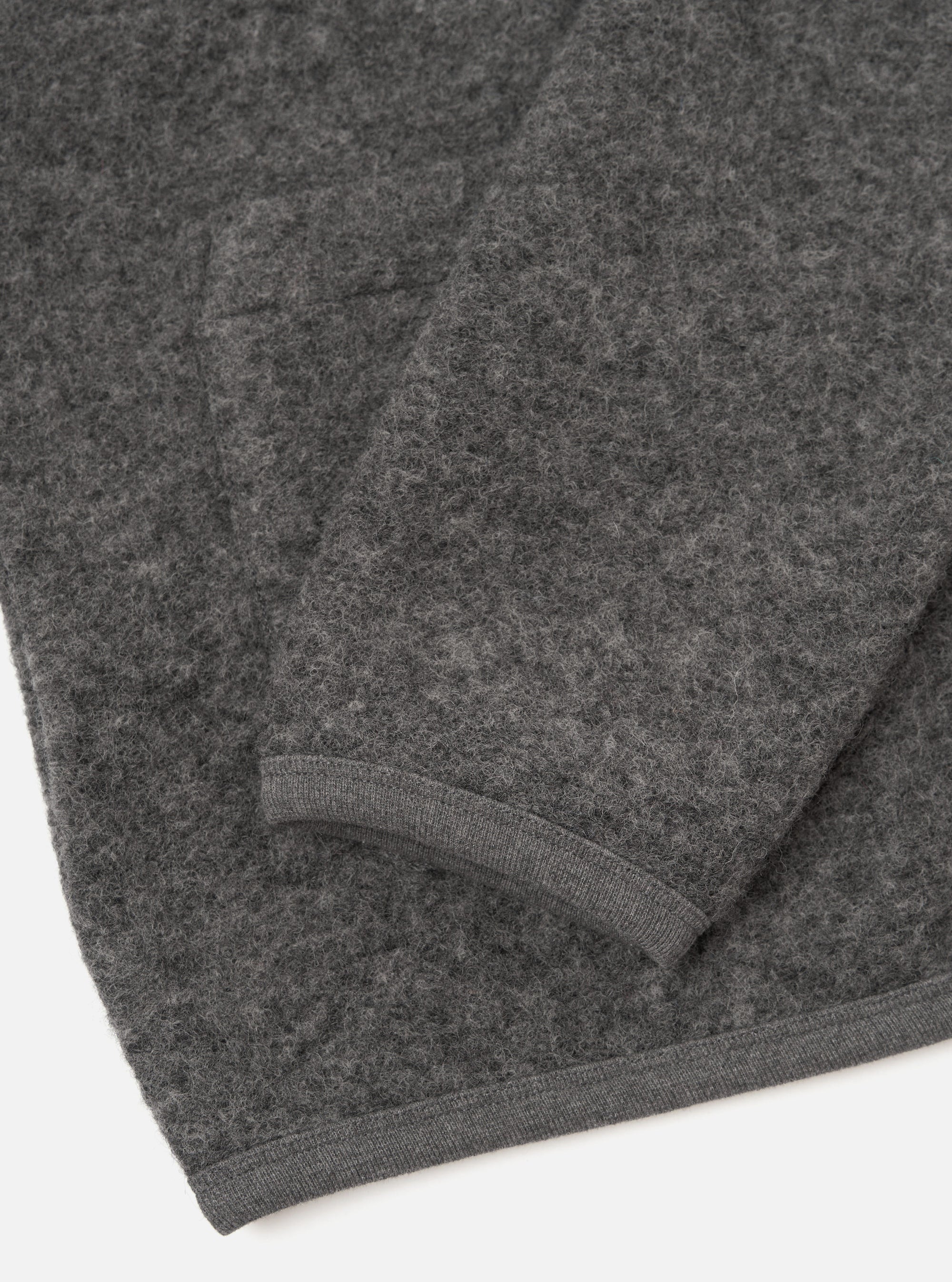 Close-up flat lay of a grey marl wool fleece cardigan on a white surface, showing the lower body and one sleeve folded back. The boiled wool texture is clearly visible across the fabric. A narrow ribbed knit hem trims the bottom edge and cuff in matching grey.