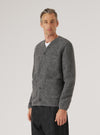Grey marl wool fleece cardigan worn buttoned by a male model on a light grey studio backdrop. V-neck, five dark horn-effect buttons down the front placket, one chest patch pocket and two lower patch pockets. Full-length sleeves with ribbed cuffs and a ribbed hem. Layered over a white T-shirt with dark trousers. Three-quarter crop, slight angle.