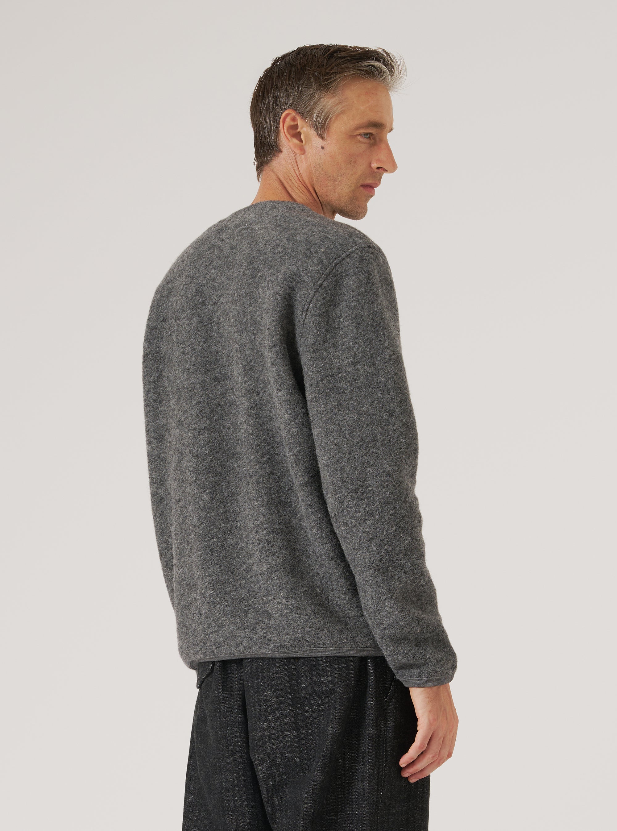 Grey marl wool fleece cardigan worn by a male model, shown from a rear three-quarter angle against a light grey studio backdrop. The brushed, textured fleece fabric has a soft, fluffy surface in a heathered grey tone. Raglan-style shoulder seams are visible. The ribbed hem and cuffs are shown. Cropped at mid-thigh, worn over dark trousers.
