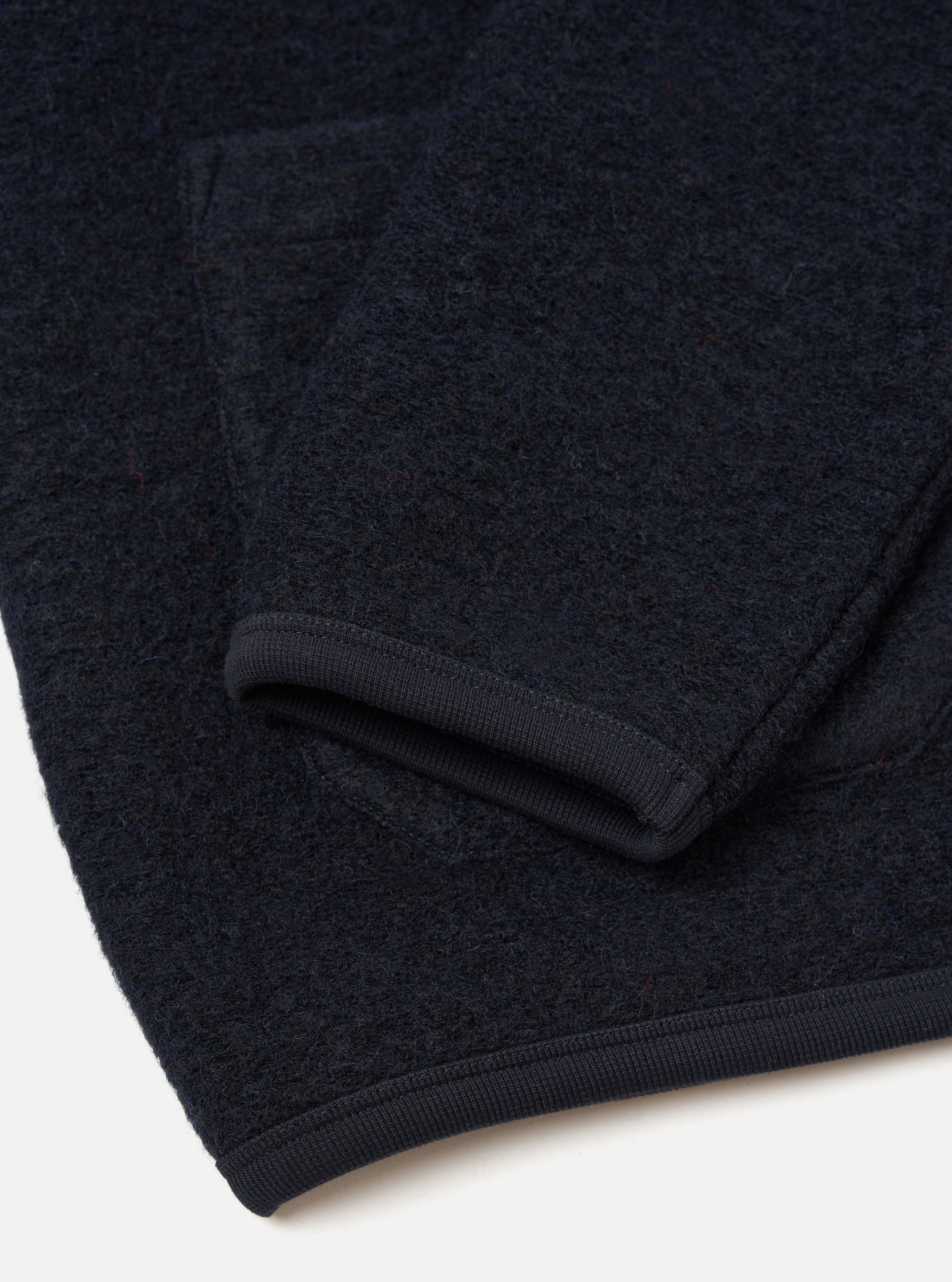 Close-up flat lay of a navy wool fleece cardigan on a white surface, showing the sleeve cuff and hem. The boiled wool texture is visible across the fabric. Both the ribbed cuff and straight hem are finished with a narrow dark navy grosgrain ribbon trim.
