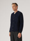 Navy wool fleece cardigan worn buttoned by a male model on a grey studio backdrop. The cardigan features a V-neck, five dark buttons down the front placket, one chest patch pocket and two lower patch pockets. The textured boiled wool fleece fabric is visible throughout. Cropped at the hip, styled over a white tee with dark grey trousers. Three-quarter front view.