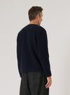 Back view of a male model wearing a navy wool fleece cardigan. The fabric has a dense, boiled-wool texture. The straight hem sits at hip level, worn untucked over dark charcoal trousers. The model turns three-quarter toward the right. Shot against a light grey studio backdrop, cropped from head to mid-thigh.