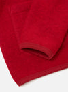 Close-up detail of a scarlet wool fleece cardigan laid flat on a white surface. The ribbed cuff is visible at the sleeve end, finished in matching red knit trim. A patch pocket is partially visible at the top of the frame. The hem also features the same ribbed trim. The fabric has a dense, textured boiled-wool fleece appearance.
