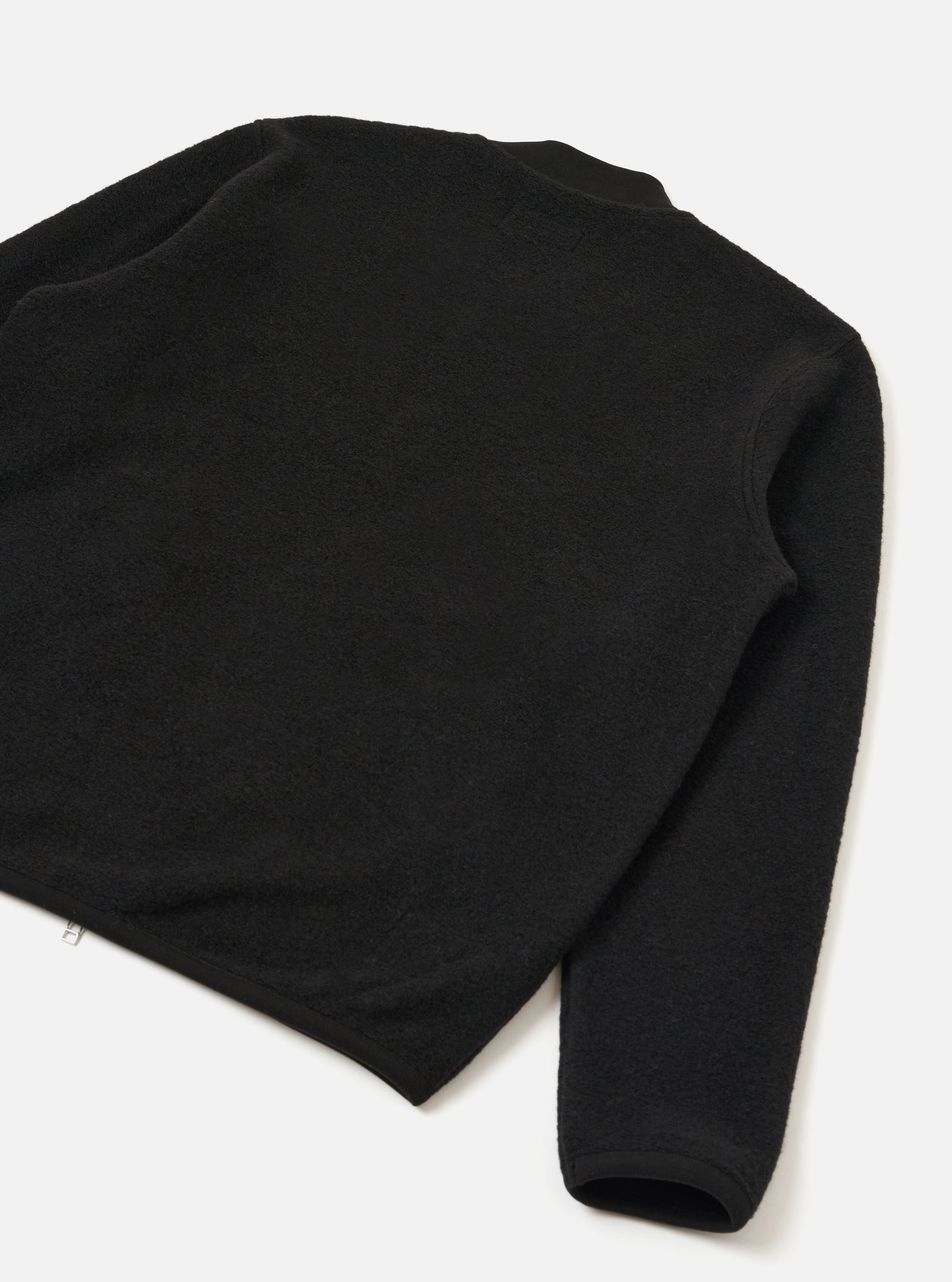 Black wool fleece zip bomber shot as a flat lay on a white surface, angled to show the back and one sleeve. The textured fleece is visible across the body and sleeve. A ribbed cuff is visible at the hem of the sleeve. A small silver zip pull is visible at the lower left hem.