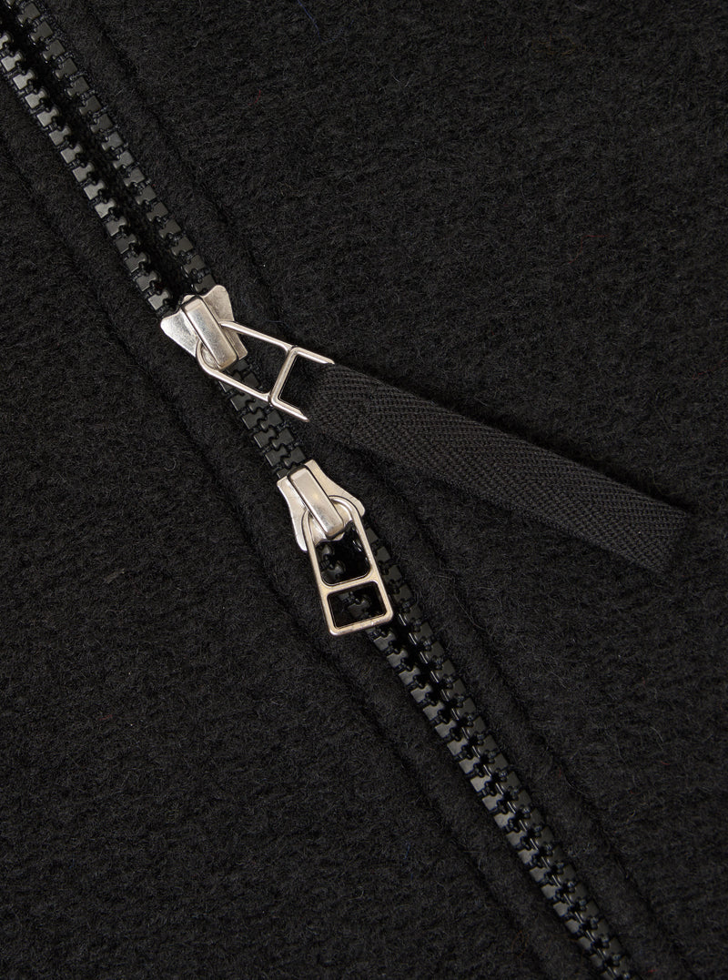 Close-up detail of the double zip fastening on a black wool fleece bomber jacket. Two silver-toned zip sliders sit on black chunky zip teeth, with the zip partially open to reveal a woven black storm placket beneath. The boiled wool fleece texture is clearly visible across the charcoal-black fabric.