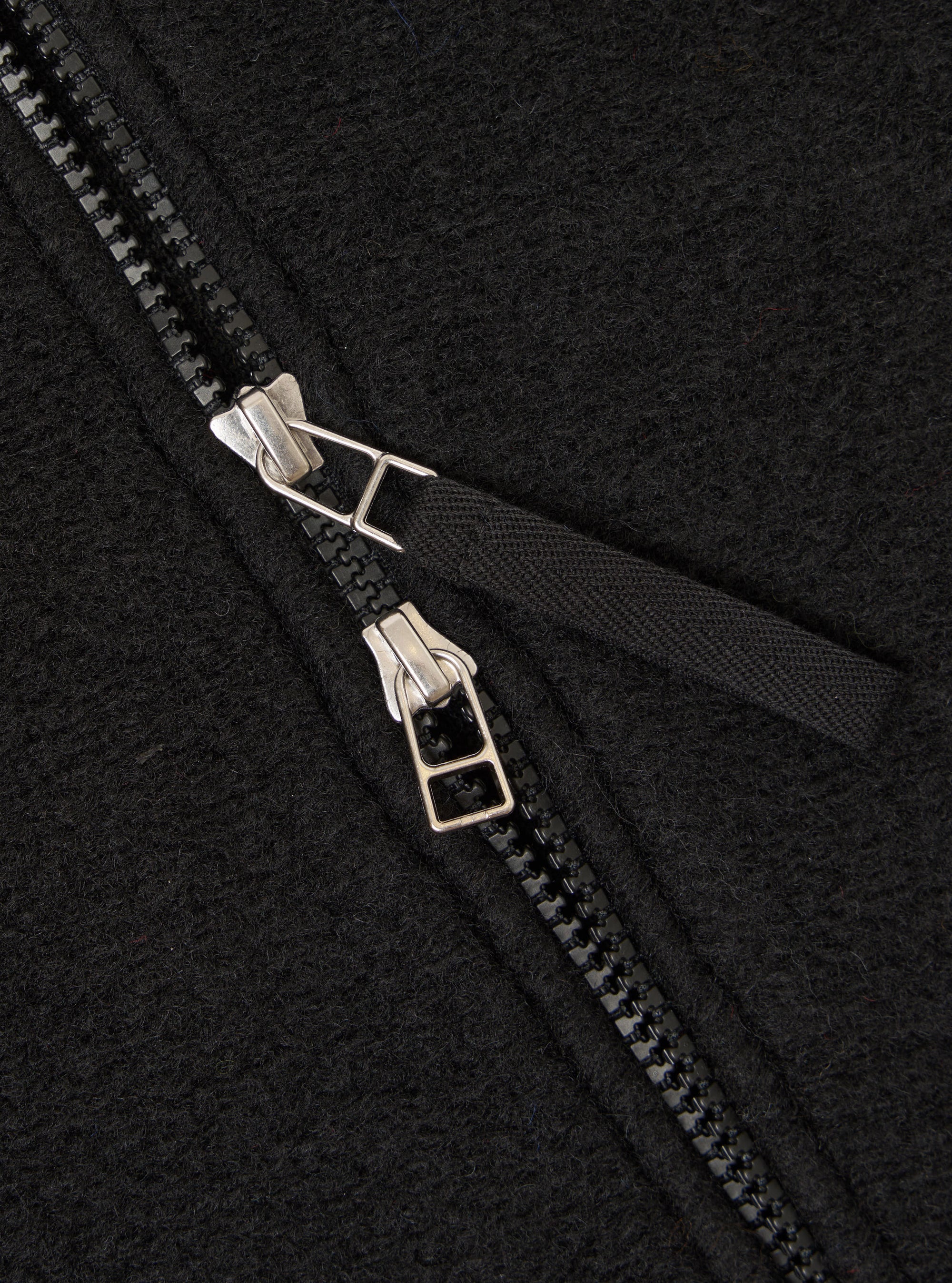 Close-up detail of the double zip fastening on a black wool fleece bomber jacket. Two silver-toned zip sliders sit on black chunky zip teeth, with the zip partially open to reveal a woven black storm placket beneath. The boiled wool fleece texture is clearly visible across the charcoal-black fabric.