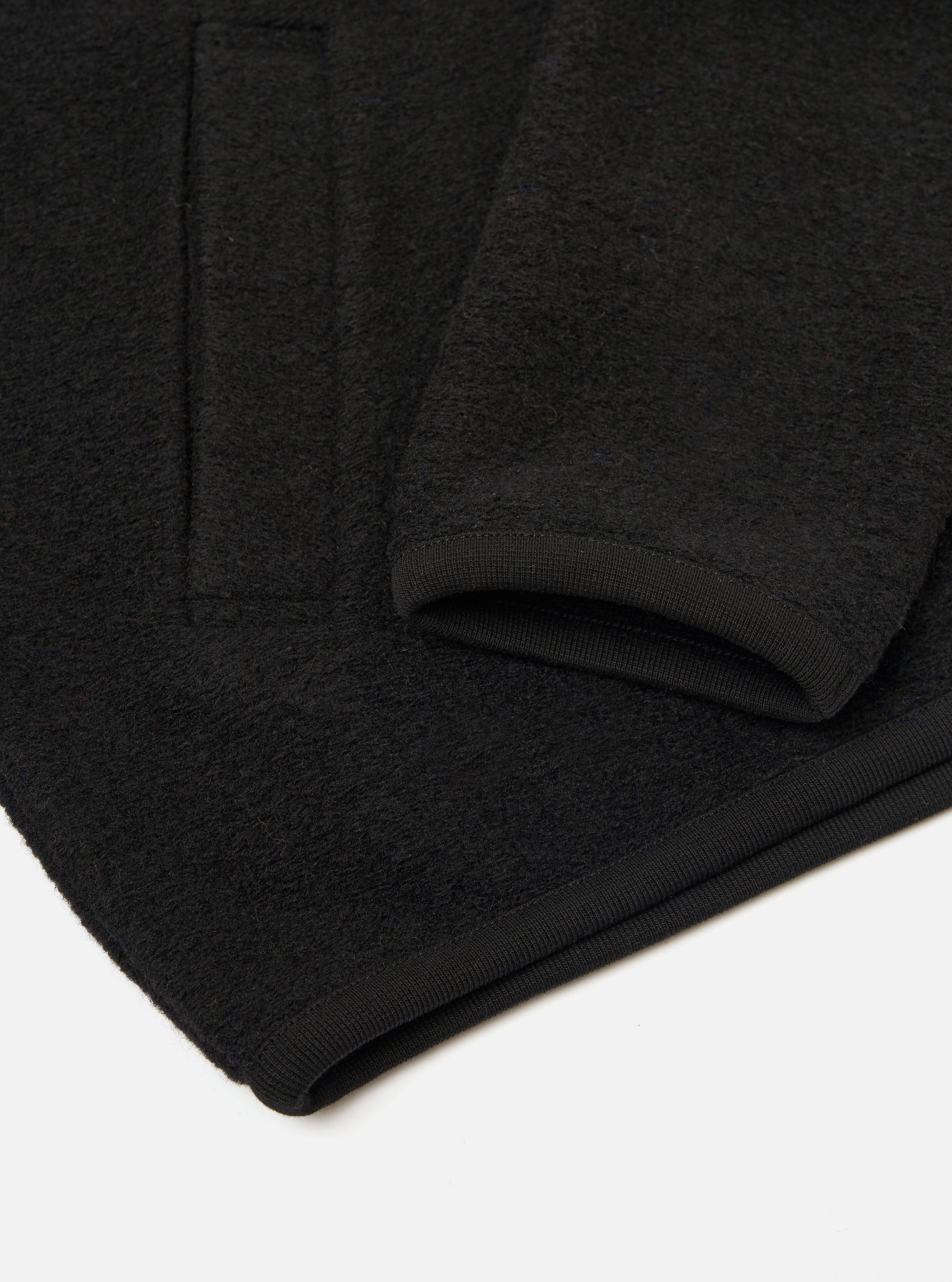 Close-up flat lay of a black wool fleece zip bomber on a white surface. The shot focuses on the lower sleeve and hem, showing the ribbed knit cuff and ribbed hem band. The dense, textured wool fleece fabric is visible in detail across the body and sleeve.