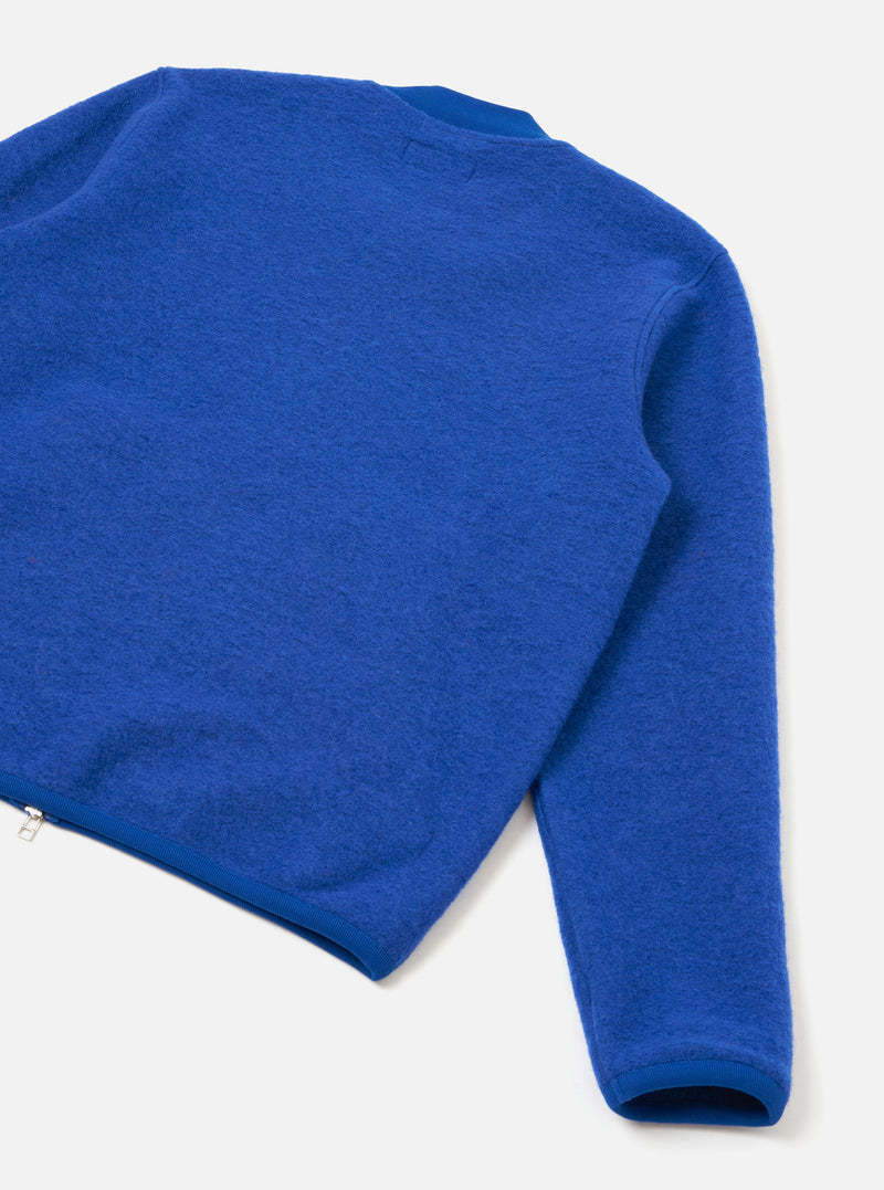Blue wool fleece zip bomber laid flat on a light grey surface, shot from above at an angle. The image shows the back and one sleeve, highlighting the brushed wool fleece texture. A silver zip is visible at the lower left hem. Ribbed cuffs and hem trim contrast with the fluffy fleece body.