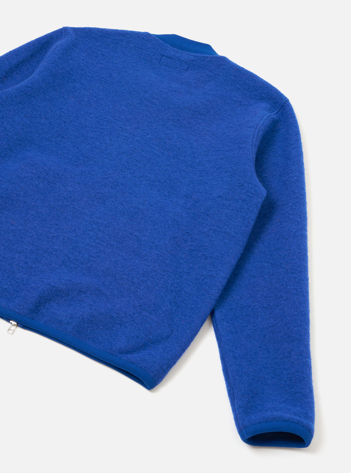 Blue wool fleece zip bomber laid flat on a light grey surface, shot from above at an angle. The image shows the back and one sleeve, highlighting the brushed wool fleece texture. A silver zip is visible at the lower left hem. Ribbed cuffs and hem trim contrast with the fluffy fleece body.