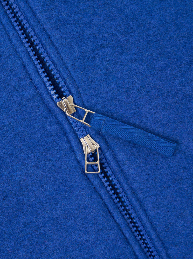 Close-up detail of the double zip fastening on a blue wool fleece zip bomber. Two gold-tone metal zip sliders sit on blue plastic teeth, with a woven blue zip pull tab. The brushed wool fleece texture is visible across the royal blue fabric.