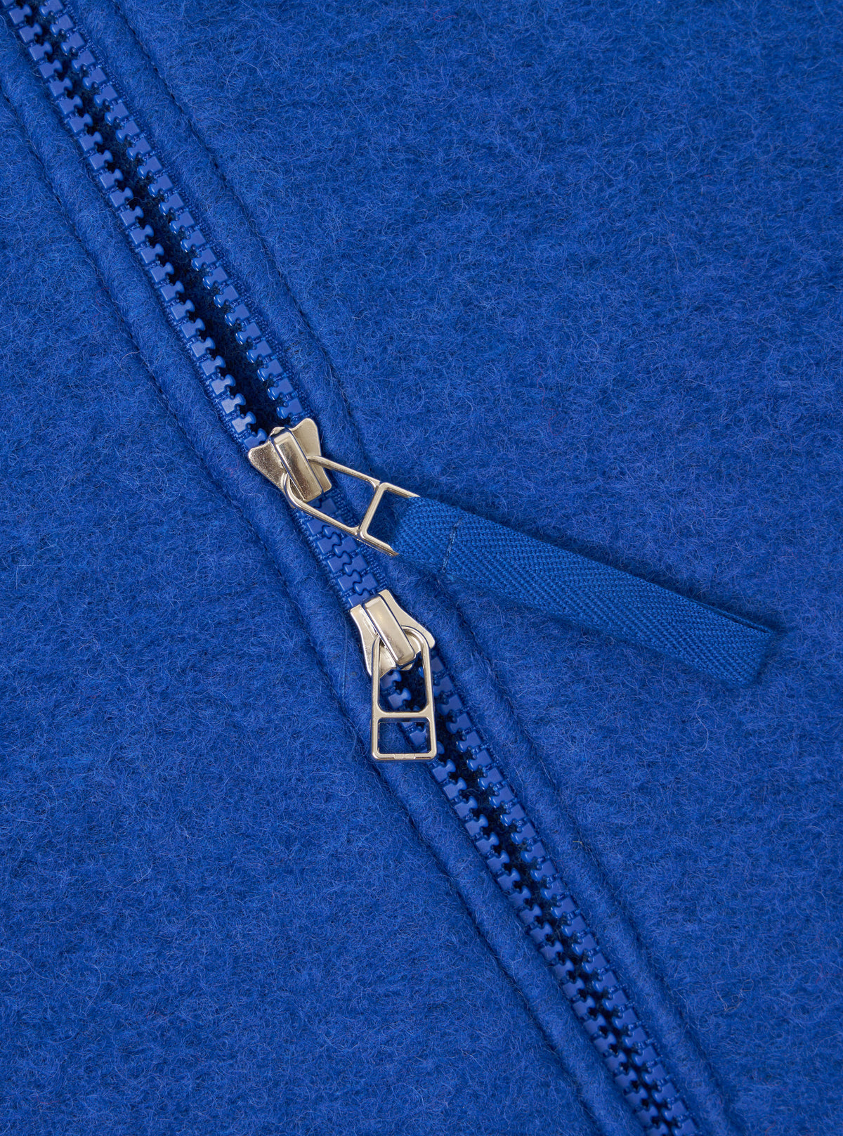 Close-up detail of the double zip fastening on a blue wool fleece zip bomber. Two gold-tone metal zip sliders sit on blue plastic teeth, with a woven blue zip pull tab. The brushed wool fleece texture is visible across the royal blue fabric.