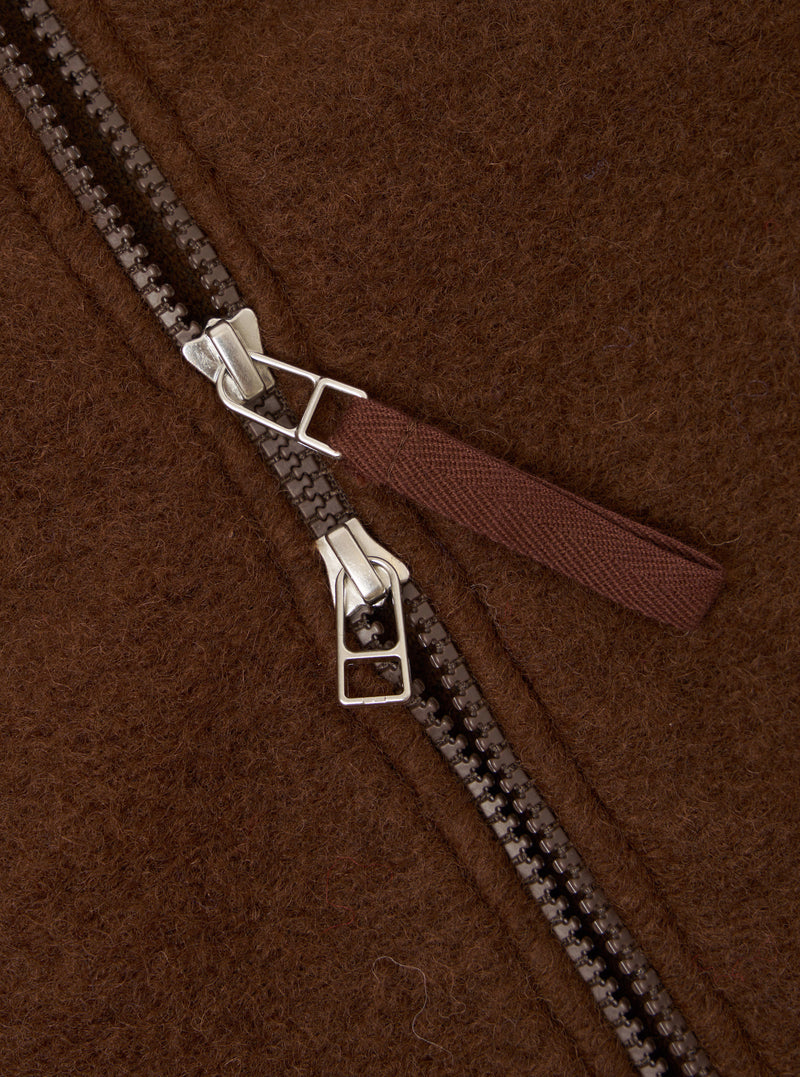 Close-up detail of the double zip on a cacao wool fleece bomber. Two gunmetal zip sliders with silver hardware sit mid-track on a dark resin chain. A woven fabric zip pull in deep burgundy-brown is attached to the upper slider. The brushed wool fleece ground shows a dense, hairy texture throughout.