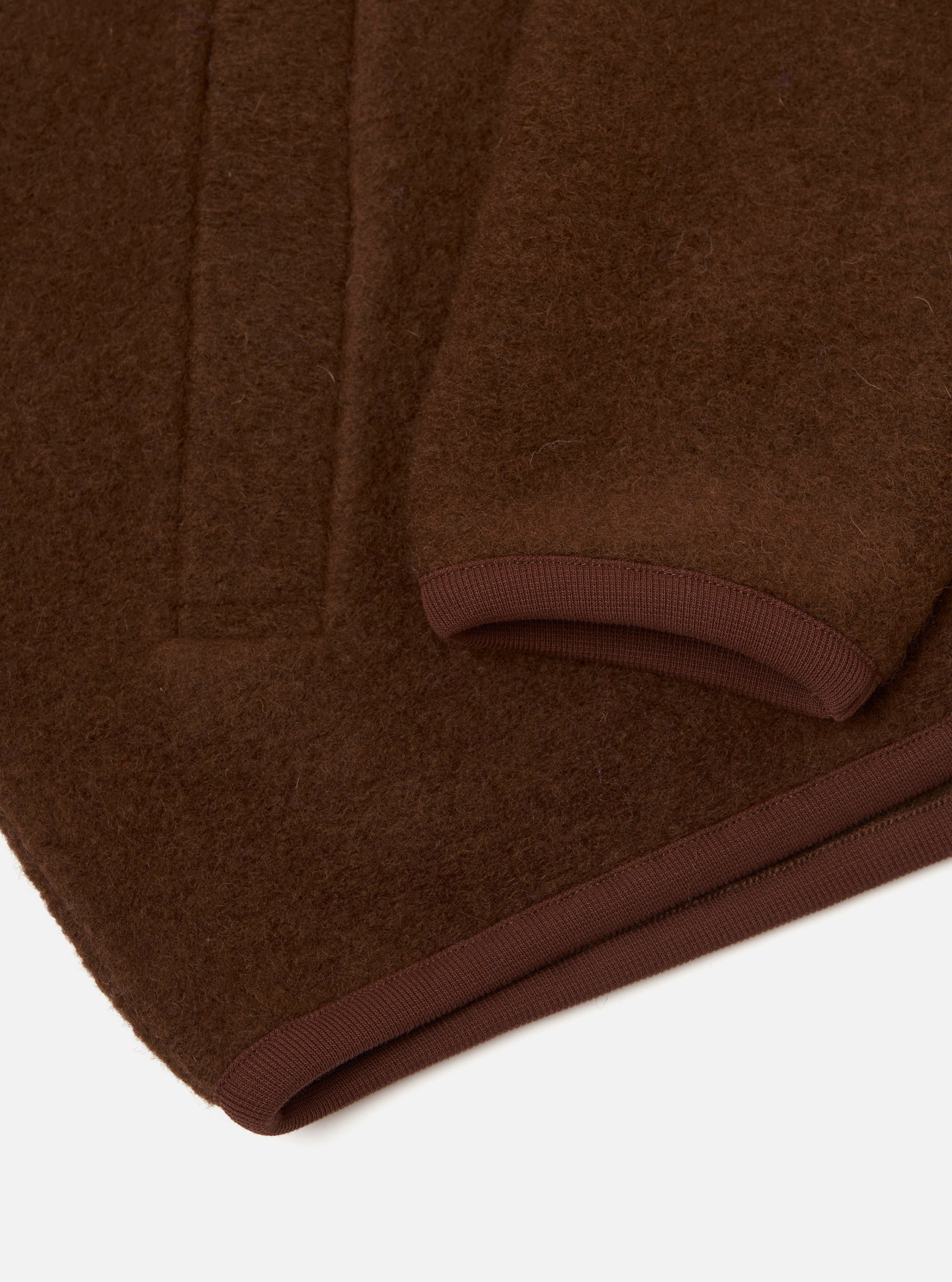 Close-up flat lay of a cacao wool fleece zip bomber on a white surface, showing the hem and sleeve cuff. The brushed wool fleece has a dense, slightly napped texture. A darker brown ribbed knit trim edges both the curved cuff opening and the straight hem. Vertical fold lines are visible across the body of the garment.
