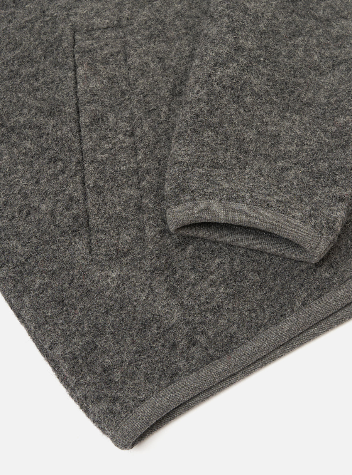 Close-up flat lay of a grey marl wool fleece zip bomber, folded to show the sleeve and hem. The boiled wool fleece has a dense, nubby texture throughout. A ribbed knit cuff is visible at the sleeve end, and a matching ribbed hem strip runs along the bottom edge. Shot on a white studio surface.