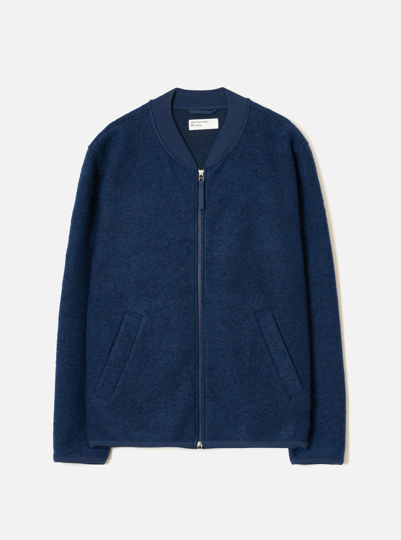 Indigo wool fleece zip bomber laid flat on a light grey studio backdrop. The jacket features a ribbed bomber collar in matching navy fabric, a full-length silver zip, two welt hip pockets, and ribbed cuffs. The boiled wool fleece has a dense, textured surface. A Universal Works woven label is visible at the neck.