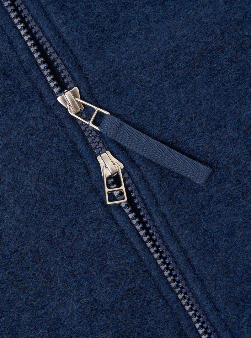 Close-up detail of the dual zip fastening on an indigo wool fleece bomber. Two silver-tone zip sliders sit together on a navy plastic-tooth zip, with a woven navy fabric pull tab. The boiled wool fleece texture is visible across the frame.