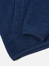 Close-up detail of an indigo wool fleece zip bomber, shot flat on a white surface. The brushed, textured fleece is visible in deep navy blue. The image focuses on the curved hem and short sleeve cuff, both finished with a smooth navy ribbed trim.