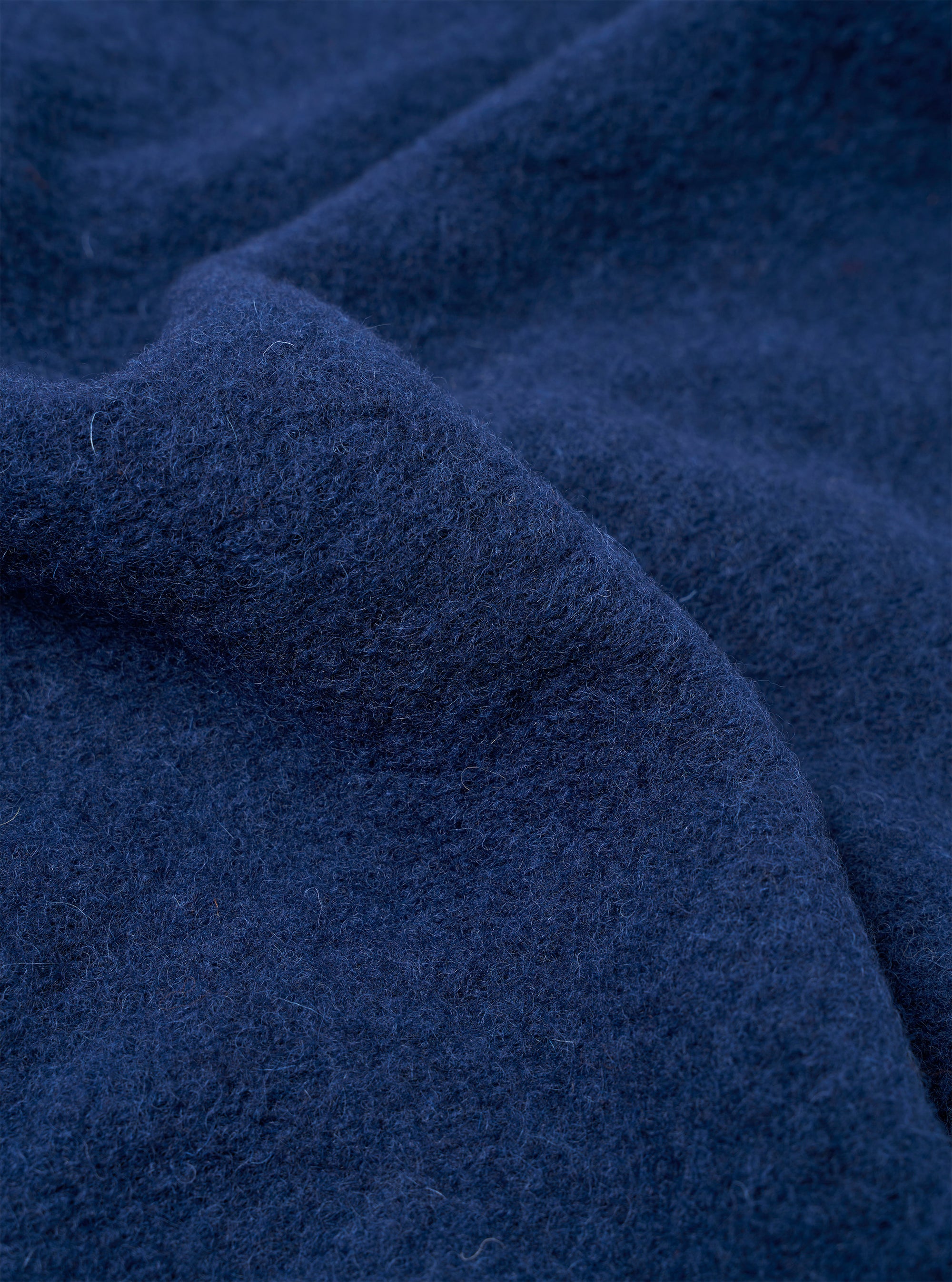 Close-up of indigo wool fleece fabric, showing a dense, brushed texture with fine curling fibres and a lightly boiled surface. The deep navy-indigo tone has subtle variation across the folds and creases of the cloth. No garment details or background are visible.