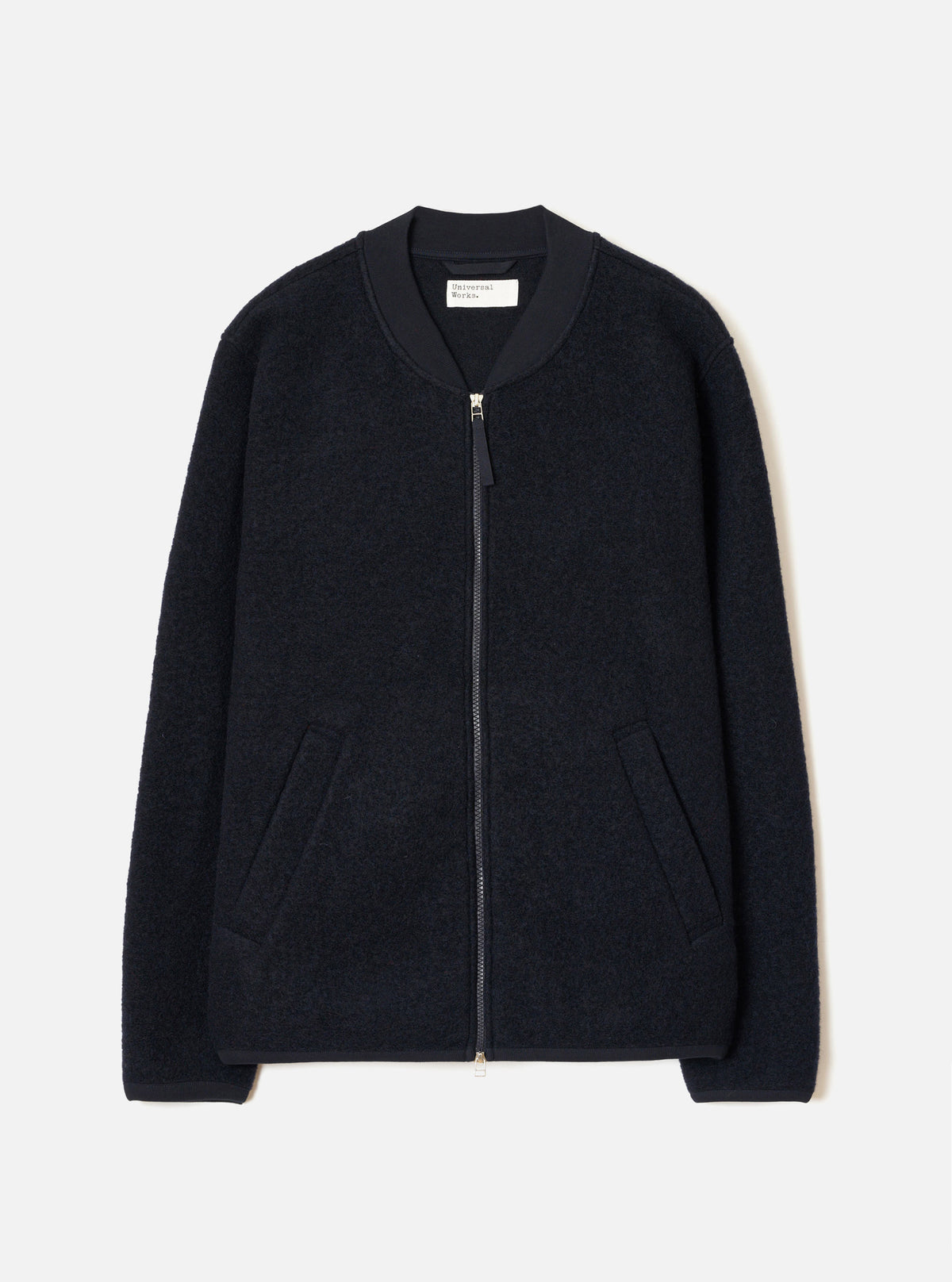 Navy wool fleece zip bomber jacket, flat lay on a light grey seamless backdrop. Textured boiled-wool-style fleece body with a smooth ribbed bomber collar in contrast navy fabric. Full-length silver zip with a small pull tab. Two welt pockets at the hip. Ribbed cuffs and hem. Universal Works woven label visible at the back neck.