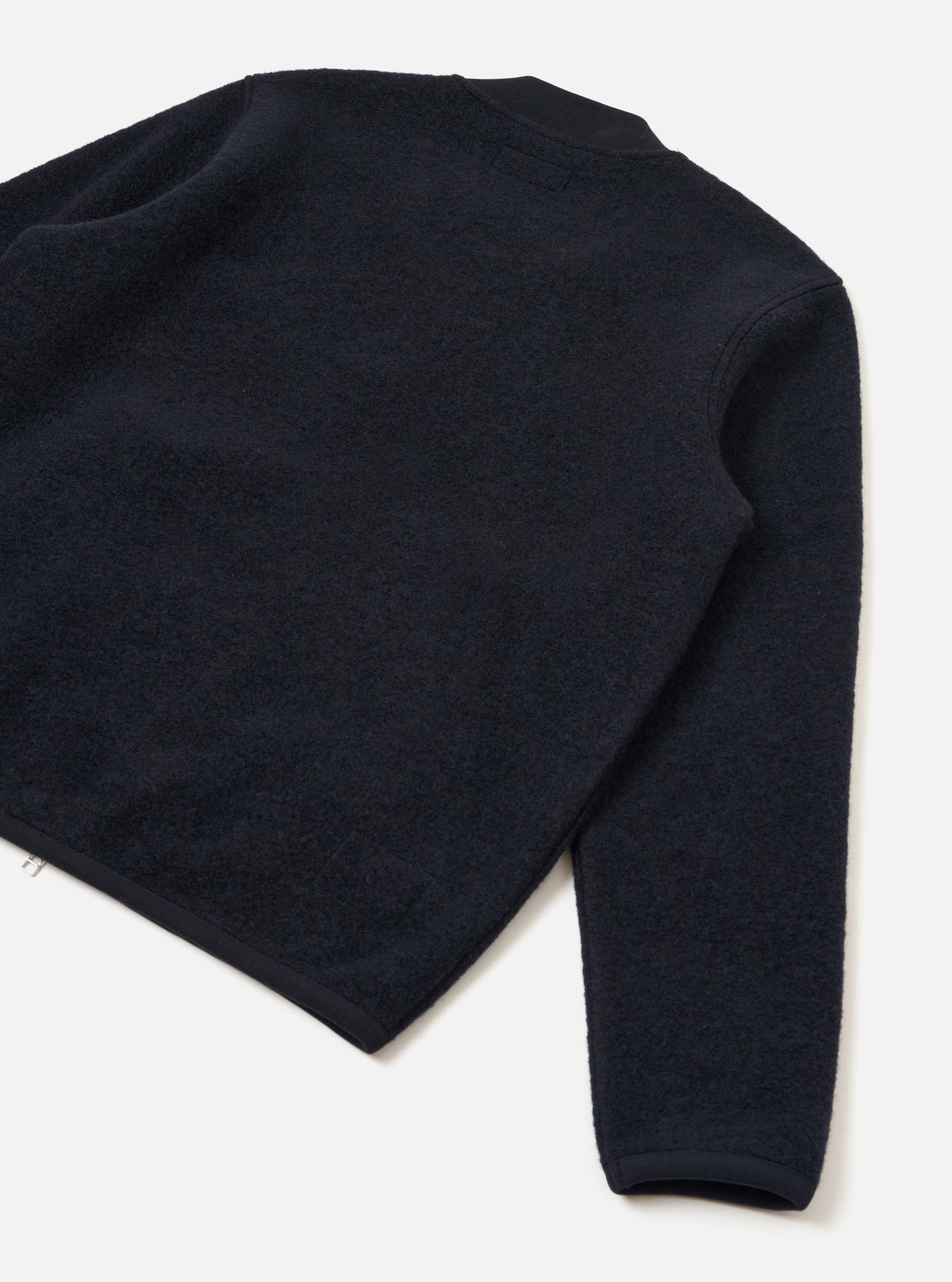 Navy wool fleece zip bomber laid flat on a white surface, shot from above at an angle showing the back and one sleeve. The textured fleece is trimmed with smooth navy grosgrain or woven binding at the crew neck, cuffs, and hem. A small silver zip pull is visible at the lower left hem.