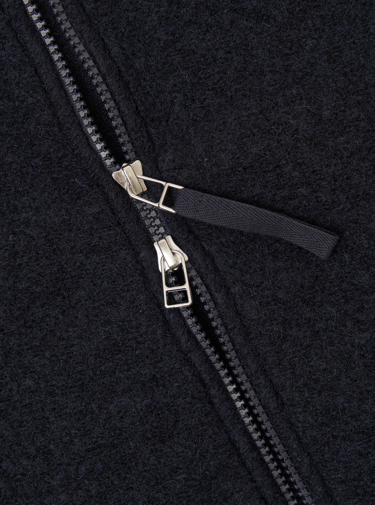 Close-up detail of the double zip fastening on a navy wool fleece bomber jacket. Two silver-tone metal zip sliders sit centrally, with black plastic teeth and a dark woven zip pull tab. The brushed wool fleece fabric texture is visible across the frame.