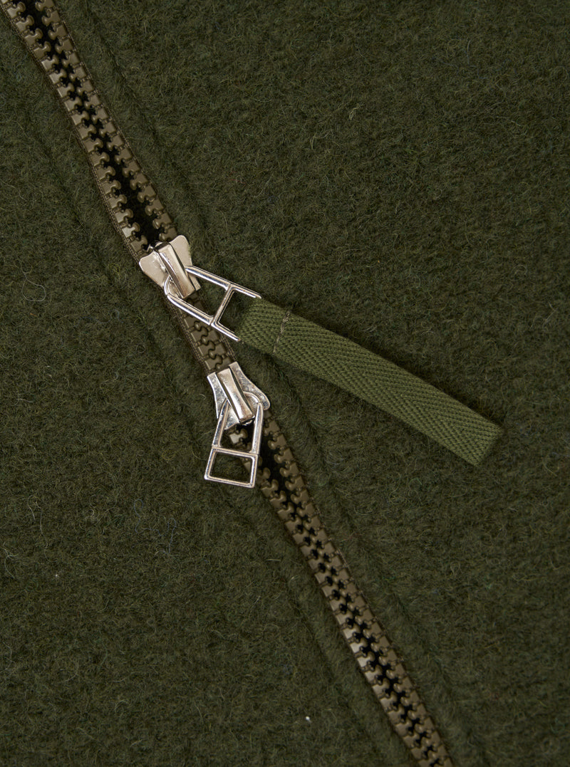 Close-up detail of the zip on an olive wool fleece bomber jacket. A chunky two-way zip with silver metal sliders and an olive woven fabric pull tab runs diagonally across the frame. The brushed wool fleece texture is visible across the background.