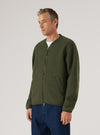 Olive wool fleece zip bomber worn by a male model on a grey studio backdrop. The jacket features a ribbed bomber collar, full-length silver zip, welt hand pockets, and ribbed cuffs. The textured boiled wool fleece is visible throughout. Worn zipped up over a white crew-neck tee, paired with dark blue jeans. Shot cropped at the thigh, three-quarter angle.