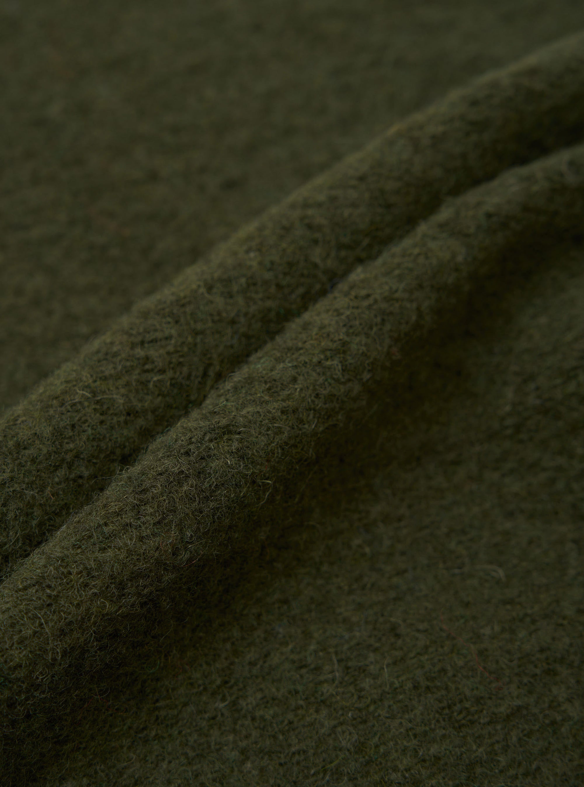 Close-up of olive wool fleece fabric showing soft, lightly brushed texture with fine surface fibres. Two rolled folds of fabric run diagonally across the frame, highlighting the fabric's weight and depth of colour. Shot against the same fabric as background.