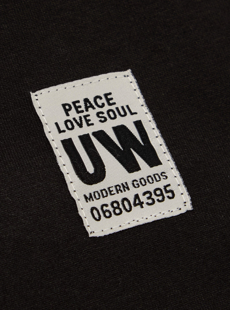 Close-up of the Black Single Jersey Core Tee showing the fabric and a tag with 'Peace Love Soul UW Modern Goods 06804395' on it. The tee is laid flat.