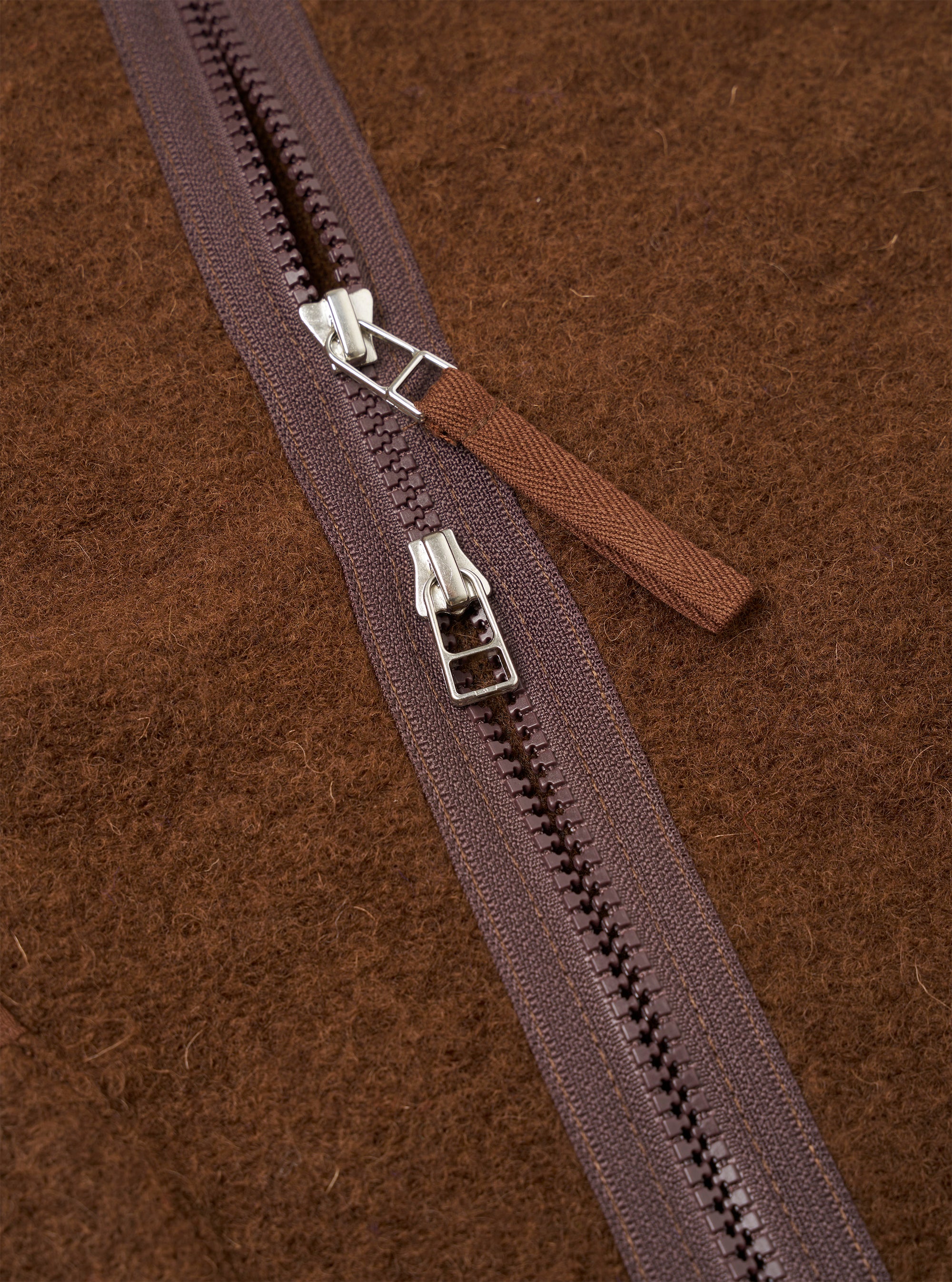 Close-up detail of the double zip fastening on a cacao wool fleece liner jacket. Two silver-hardware zip sliders sit on a dark plum zip tape with moulded plastic teeth. A woven brown zip garage tab is visible. The brushed, textured wool fleece fabric fills the background.