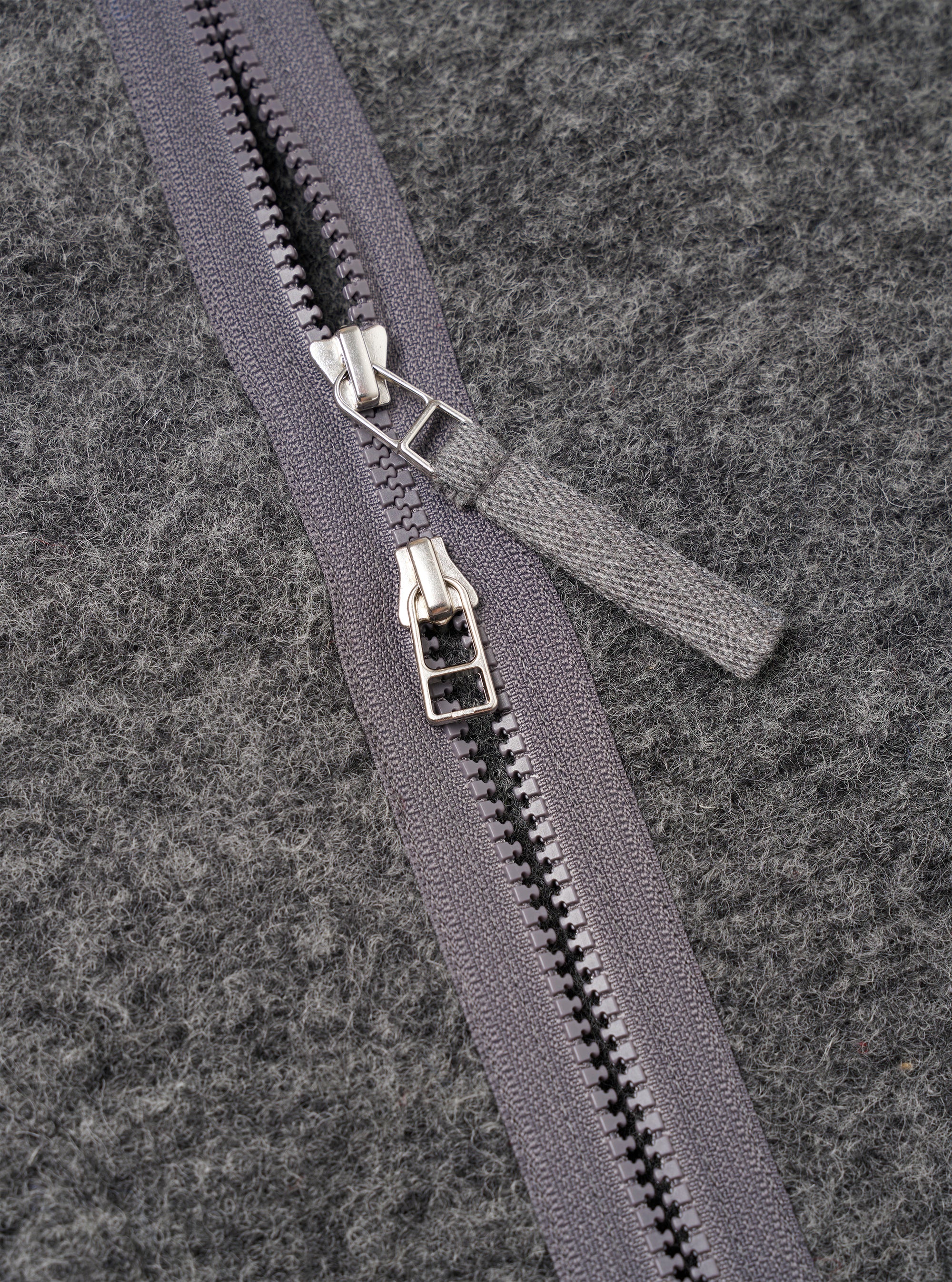 Close-up detail of the grey marl wool fleece zip liner jacket showing a two-way centre-front zip with two silver metal sliders and a fabric pull tab in matching grey. The zip tape is dark grey with black plastic teeth. The brushed wool fleece fabric texture is visible across the background.
