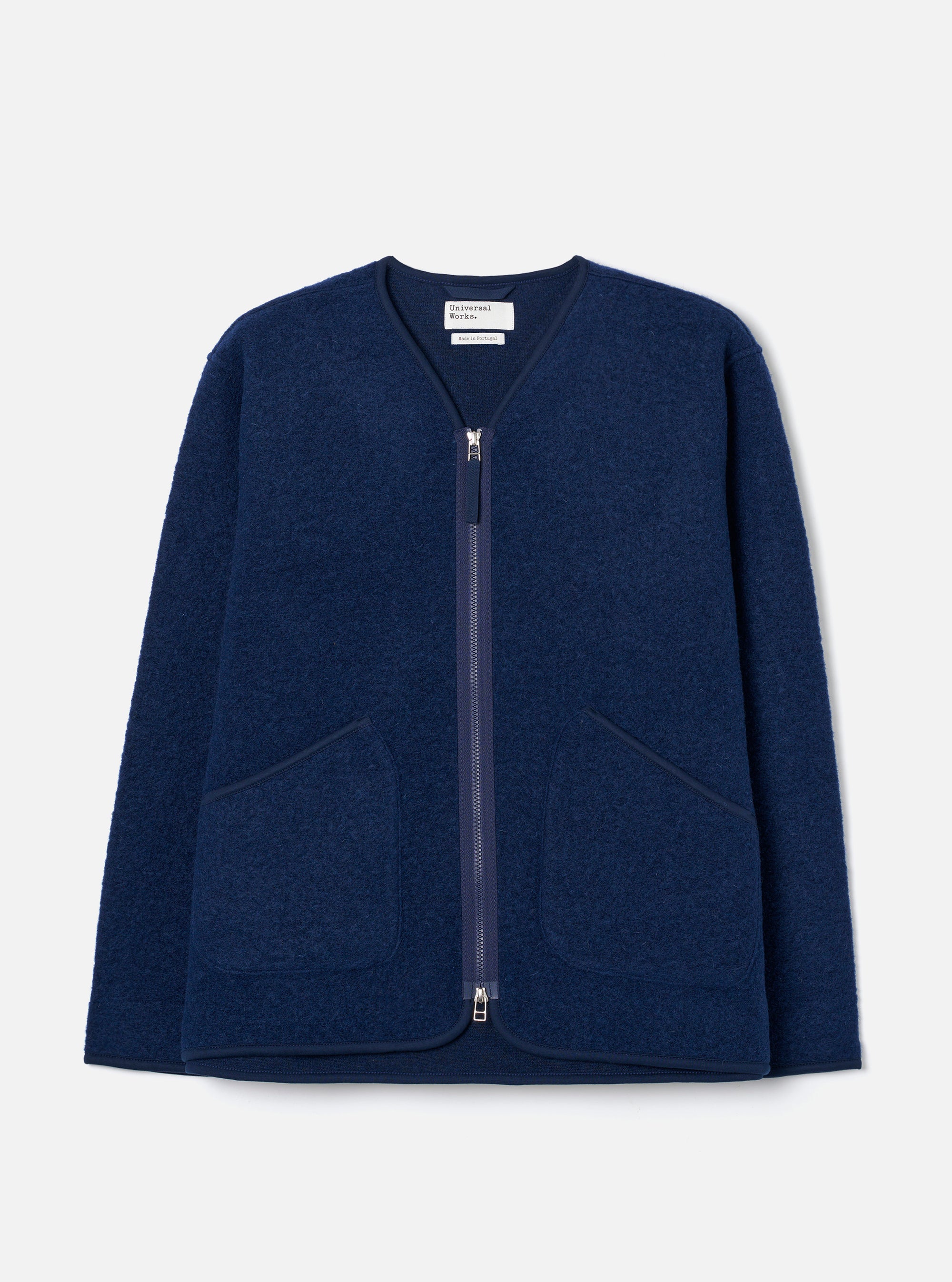 Indigo wool fleece zip liner jacket laid flat on a light grey seamless backdrop. Collarless V-neckline with navy binding trim, full-length silver zip, and two large patch pockets with matching bound edges. Textured boiled wool fleece fabric throughout. Universal Works woven label visible at the neck.
