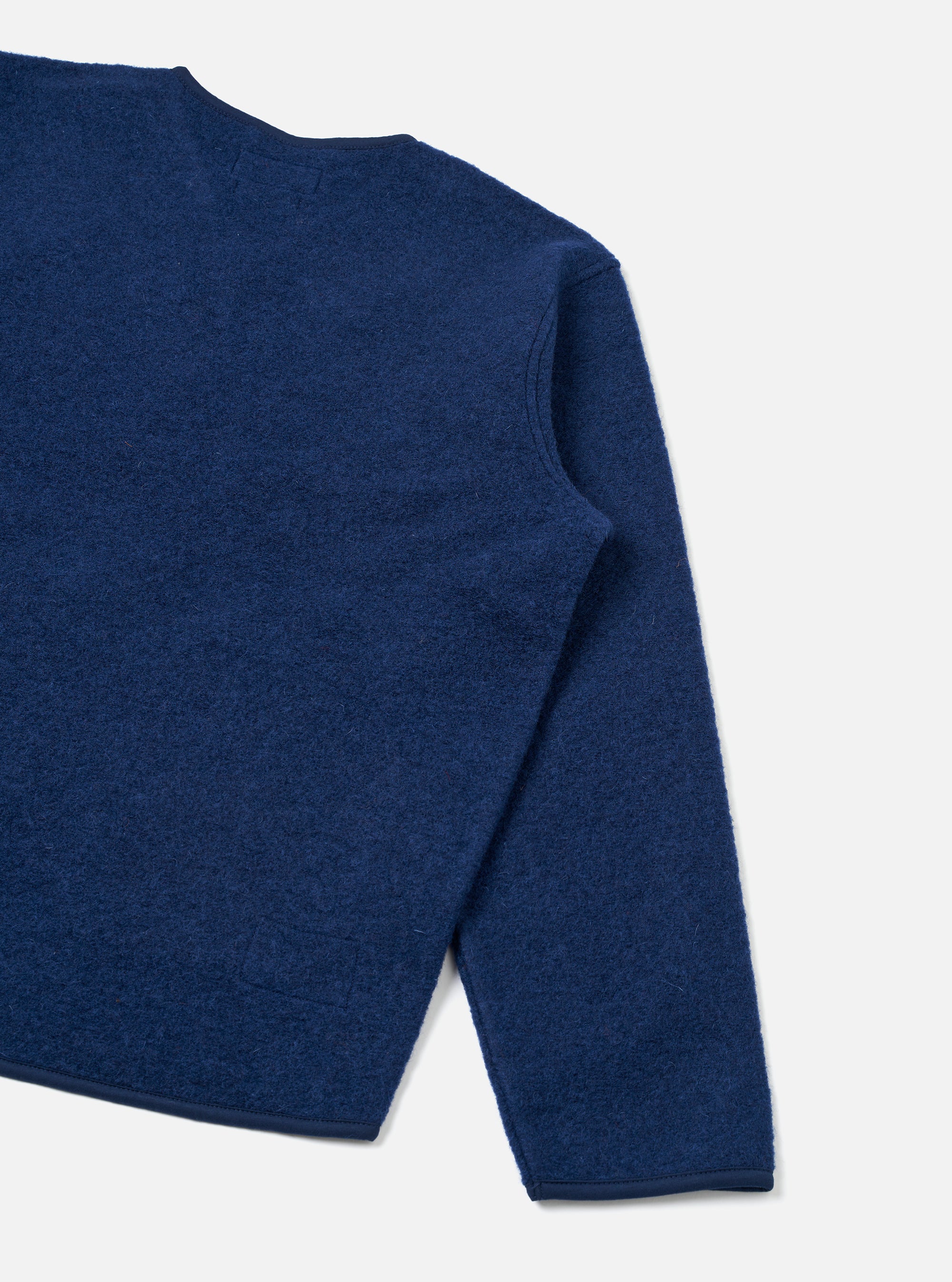 Close-up of an indigo wool fleece zip liner jacket on a plain light grey studio backdrop. The shot shows the upper chest, one shoulder and a full sleeve, cropped at the hem. The boiled wool fleece texture is visible across the body and sleeve, with a narrow bound cuff edge and a patch pocket partially visible at the lower left.