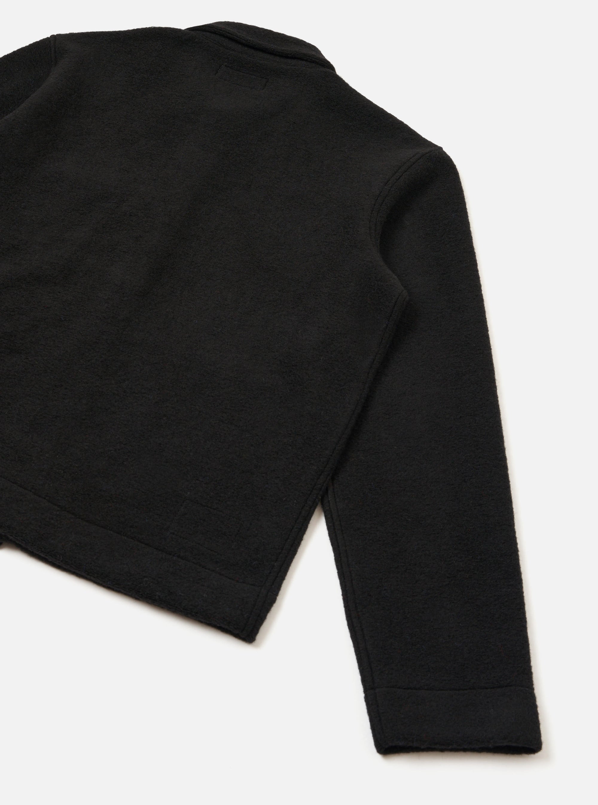 Close-up flat lay of a black wool fleece uniform jacket on a white studio surface, showing the back and one sleeve. The textured fleece fabric is visible across the body and sleeve. A ribbed cuff finishes the sleeve hem, and a wide ribbed band runs along the jacket's lower hem.