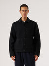 Black wool fleece uniform jacket worn buttoned by a male model against a light grey studio backdrop. The jacket features a spread collar, five dark buttons down the centre front, a chest patch pocket, and two large lower patch pockets. The textured boiled wool fleece fabric is visible throughout. Cropped at the hip, layered over a white T-shirt and navy trousers.