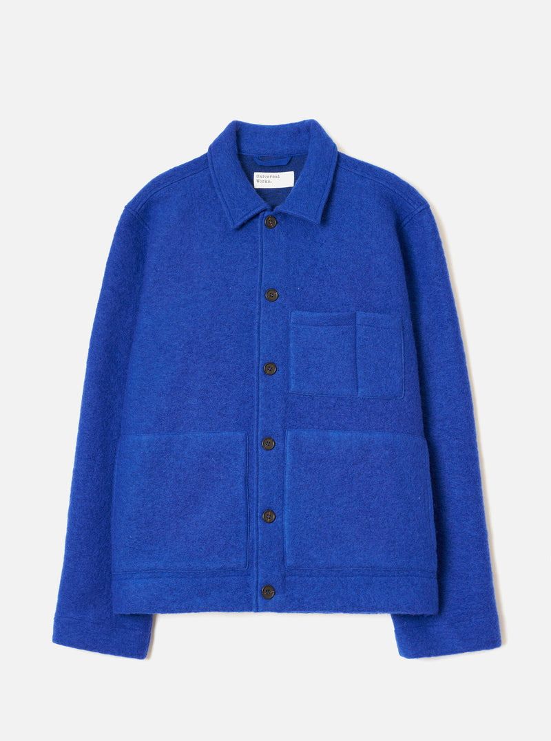 Blue wool fleece uniform jacket laid flat on a light grey studio backdrop. Features a point collar with a Universal Works label, a five-button front placket with dark brown buttons, one chest patch pocket, two large lower patch pockets, and ribbed cuffs. The boiled wool texture is visible throughout.