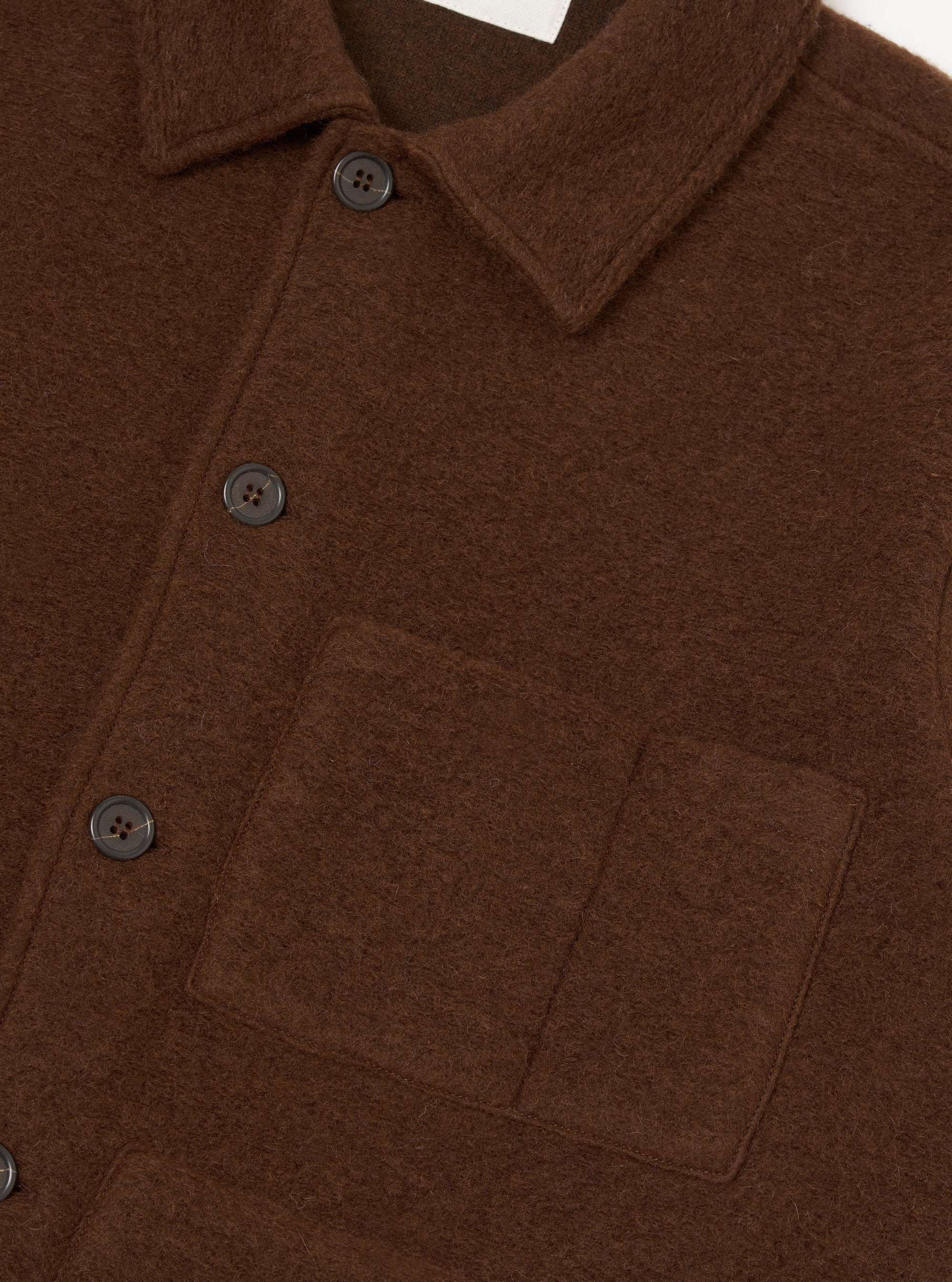 Close-up of a cacao wool fleece uniform jacket on a plain white backdrop. The shot shows the spread collar, button placket with dark four-hole buttons, and a patch pocket on the chest. The brushed wool fleece texture is clearly visible across the body.