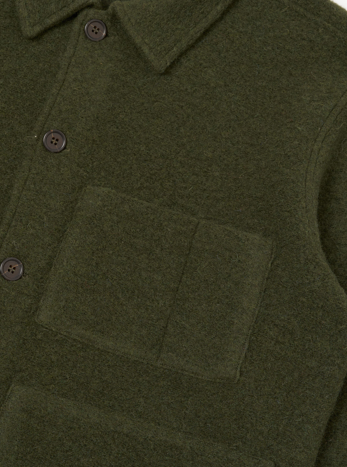 Close-up of an olive wool fleece uniform jacket on a plain white studio backdrop. The shot shows the point collar, button placket with dark four-hole buttons, and a large patch pocket on the chest. The boiled wool texture is clearly visible throughout.