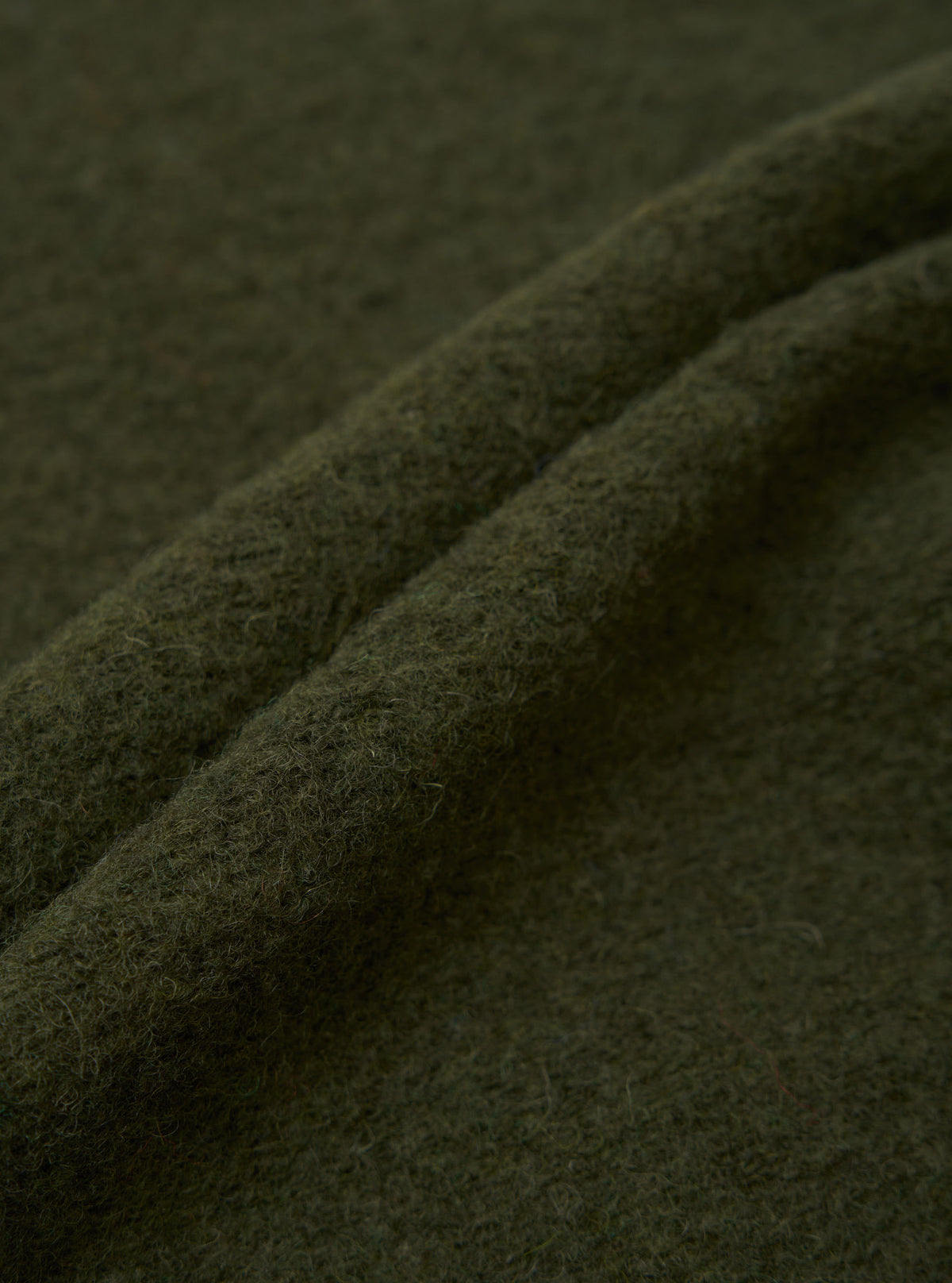 Close-up of olive wool fleece fabric showing soft, brushed texture with fine surface fibres. Two rounded folds run diagonally across the frame, highlighting the fabric's depth and weight. Solid khaki-green colour throughout, photographed flat against itself with no background visible.