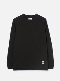 Black long-sleeve jersey T-shirt laid flat on a light grey studio backdrop. Crew neck with a visible Universal Works woven label at the back neck. Single chest patch pocket on the left breast. Ribbed cuffs at the wrists. Small branded UW woven badge at the lower right hem. Plain smooth fabric throughout.