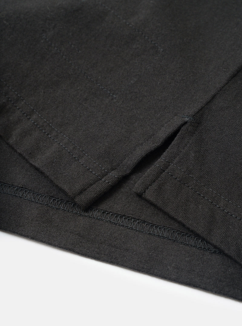 Close-up detail of a black long-sleeve pocket tee in single jersey cotton, flat lay on a white surface. The shot focuses on the lower hem and side seam vent, showing zigzag overlock stitching along the hem edge and a small side split. The jersey fabric has a fine, slightly textured weave visible throughout.