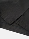 Close-up detail of a black long-sleeve pocket tee in single jersey cotton, flat lay on a white surface. The shot focuses on the lower hem and side seam vent, showing zigzag overlock stitching along the hem edge and a small side split. The jersey fabric has a fine, slightly textured weave visible throughout.