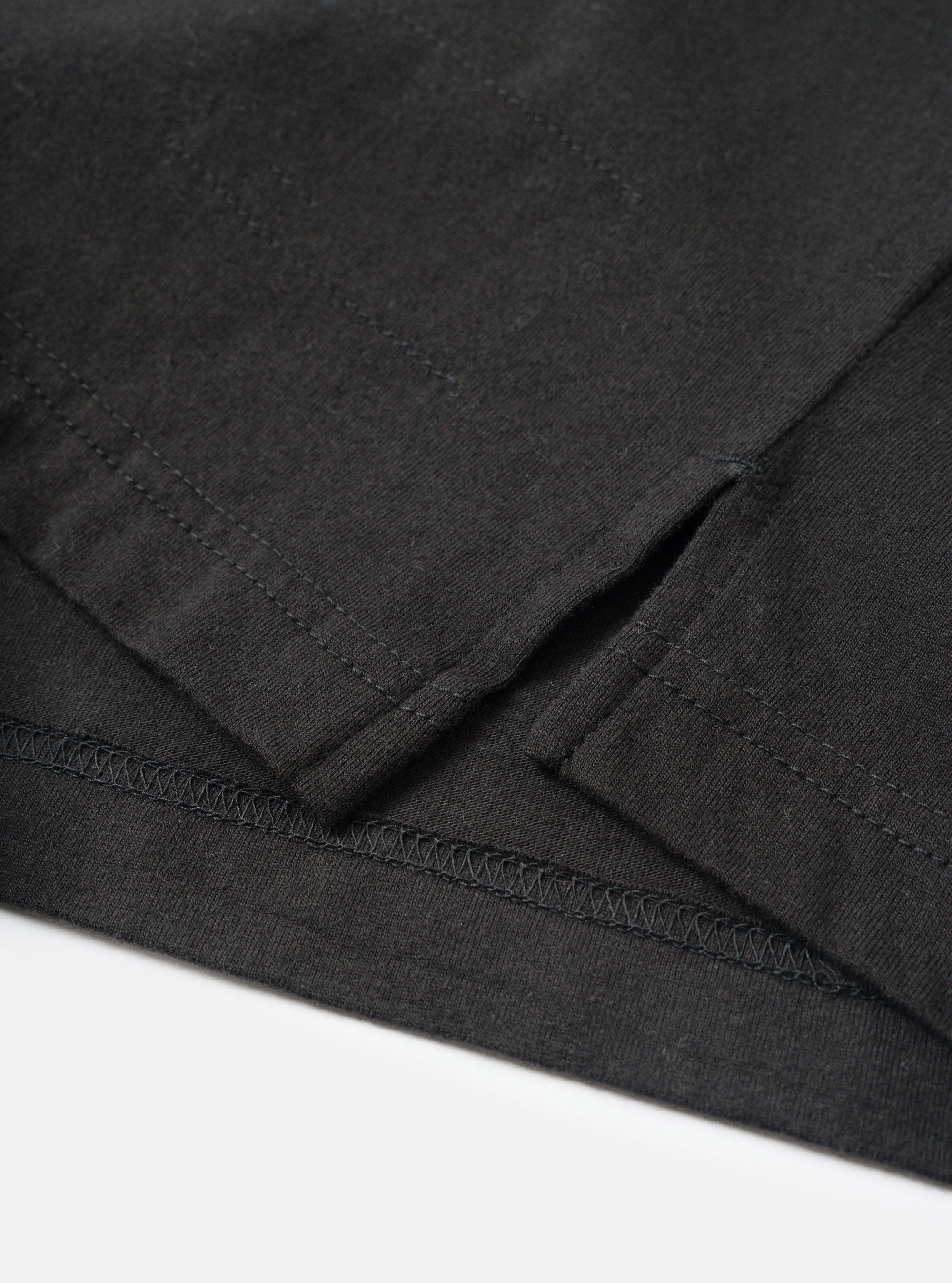 Close-up detail of a black long-sleeve pocket tee in single jersey cotton, flat lay on a white surface. The shot focuses on the lower hem and side seam vent, showing zigzag overlock stitching along the hem edge and a small side split. The jersey fabric has a fine, slightly textured weave visible throughout.