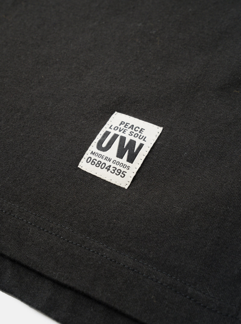 Close-up detail shot of a black long-sleeve pocket tee in single jersey cotton. A rectangular woven label reading "Peace Love Soul UW Modern Goods 06804395" is stitched onto the chest pocket. The hem and pocket stitching are visible. Shot as a flat lay on a white surface.