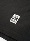 Close-up detail shot of a black long-sleeve pocket tee in single jersey cotton. A rectangular woven label reading "Peace Love Soul UW Modern Goods 06804395" is stitched onto the chest pocket. The hem and pocket stitching are visible. Shot as a flat lay on a white surface.